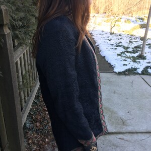 May include: A black quilted jacket with a green and red trim. The jacket has a textured surface and a long, fitted silhouette.