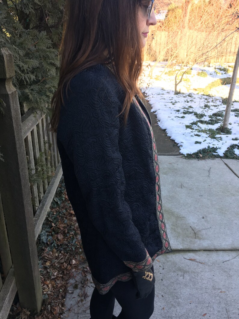 May include: A black quilted jacket with a green and red trim. The jacket has a textured surface and a long, fitted silhouette.