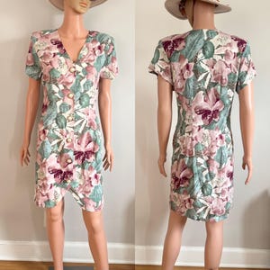 May include: A vintage floral print dress featuring a V-neckline and short sleeves. The dress showcases a pattern of pink and purple flowers with green leaves on a cream background. It has gold-colored buttons down the front and a knee-length hemline.