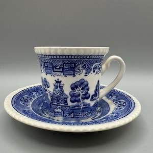 May include: A blue and white teacup and saucer set. The cup and saucer feature a detailed blue Willow pattern design on a white background. The cup has a curved handle and a scalloped rim. The saucer has a matching design and a similar scalloped edge.