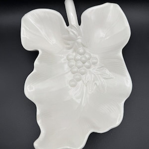 May include: A white ceramic serving dish shaped like a grape leaf. It has a raised design of grapes and leaf veins. The handle is a curved stem. The dish is set against a black background.