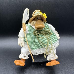 May include: A whimsical duck figurine wearing a straw hat with a yellow flower, a green and white checkered dress, and white lace-trimmed pants. The duck holds a fishing net and has orange feet. The figurine is on a black surface.