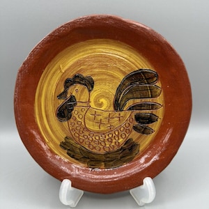 May include: A round, hand-painted ceramic plate with a rooster design. The plate features a brown rim and a yellow and brown center. The rooster is depicted in brown and black, with a detailed tail and body.