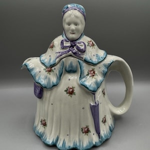 May include: A ceramic teapot in the shape of a woman, featuring a white dress adorned with small pink flowers and blue trim. The teapot has a handle and a spout, and the woman's head forms the lid. A purple bow and umbrella are also visible.