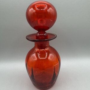 Może przedstawiać: Wintage czerwona szklana karafka z okrągłym korkiem. Karafka ma pękaty korpus z pionowymi żebrami i rozszerzoną podstawą. Korek to duża, okrągła kula. Szkło jest głęboko, przezroczysto czerwone.
