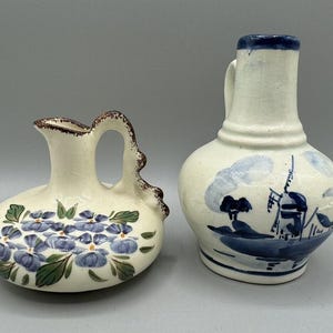 May include: Two ceramic pitchers with cream-colored glazes. The left pitcher features a low, rounded body with hand-painted blue flowers and green leaves. The right pitcher is taller with a narrow neck and blue painted windmill scene.