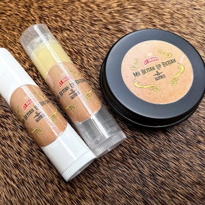 Puede incluir: Un surtido de productos para el cuidado de los labios, que incluye dos bálsamos labiales y una pequeña lata redonda. Los bálsamos labiales están en tubos transparentes con etiquetas marrones, y la lata tiene una etiqueta marrón con letras doradas. El texto de las etiquetas dice "Mo Bettah Lip Buttah".