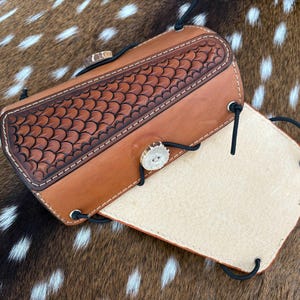 May include: A handcrafted leather case in shades of brown, featuring a detailed embossed pattern on the top flap. The case is secured with a button made from a natural material and black cords. The interior is a lighter tan color.