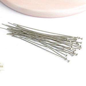 May include: A bunch of silver head pins with round heads. The pins are arranged in a loose pile on a white surface.