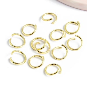 Puede incluir: Primer plano de un grupo de 10 anillos de salto dorados. Los anillos están dispersos sobre una superficie blanca.