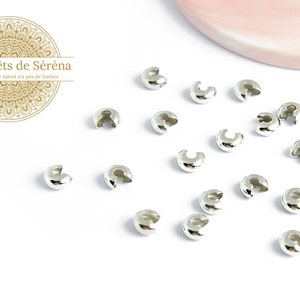 Peut inclure: Gros plan sur des perles à sertir de couleur argentée, éparpillées sur une surface blanche. Les perles sont petites et rondes, avec une petite ouverture sur un côté.