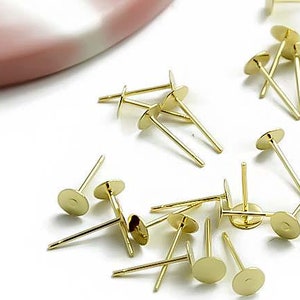 Peut inclure: Gros plan sur un groupe de tiges de boucles d'oreilles dorées avec des sommets ronds plats. Les tiges sont dispersées sur une surface blanche.