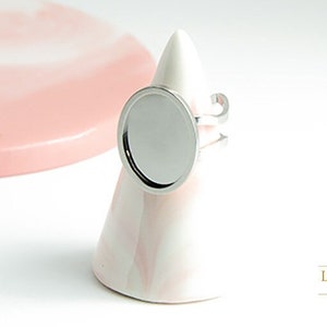 Peut inclure: Une bague en argent avec un cabochon rond, vide, de couleur argentée. La bague est sur un support conique blanc et rose.