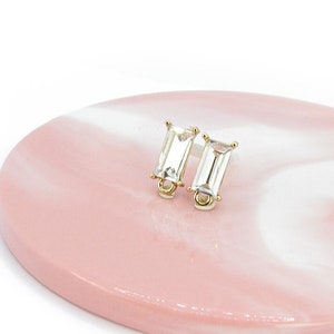 Może przedstawiać: Para złotych kolczyków z przezroczystymi prostokątnymi kamieniami. Kolczyki leżą na różowo-białym marmurowym podłożu.