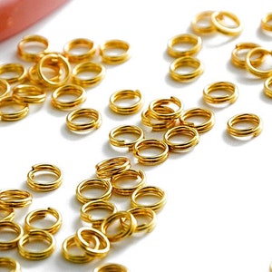 Op de afbeelding: Een grote hoeveelheid kleine, goudkleurige springringen. De ringen zijn verspreid over een wit oppervlak.