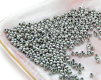 May include: A close-up of a small, white square dish with a gold rim. The dish is filled with a large quantity of small, silver-colored metal beads.