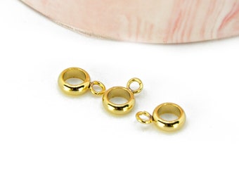 May include: Three gold-coloured jewellery findings with small loops, arranged on a white surface. The findings are circular with a small loop attached. The image also includes a logo with the text "Les Apprêts de Séréna".