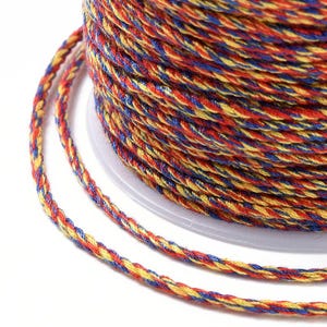 May include: Close-up of a spool of colourful twisted cord. The cord is a mix of red, yellow, and blue threads, tightly wound together. The cord is on a white spool, and the image is well-lit.