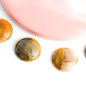 Peut inclure: Quatre pierres rondes et polies, dans des tons de marron, de jaune et de gris, sont disposées sur une surface blanche. Une forme rose et incurvée est en arrière-plan. Les pierres ont un aspect marbré, suggérant des matériaux naturels.