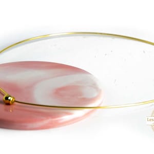 Op de afbeelding: Een gouden choker ketting met een kleine ronde kraal. De ketting wordt getoond op een roze en wit marmeren oppervlak.