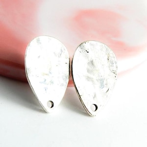 Könnte beinhalten: Zwei silberfarbene, tropfenförmige Ohrringe mit gehämmerten Texturen. Jeder Ohrring hat unten ein kleines Loch. Die Ohrringe werden auf einer weißen Oberfläche mit einem rosa-weißen Hintergrund präsentiert.