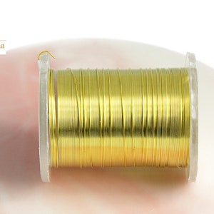 Op de afbeelding: Een spoel goudkleurig draad, strak gewikkeld op een transparante plastic spoel. De draad heeft een heldere, metallic goudkleur. De spoel staat tegen een zachte, roze achtergrond. Het logo "Les Apprêts de Séréna" is zichtbaar.