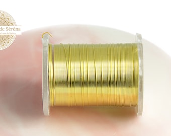 Pode incluir: Uma bobina de fio dourado, enrolada firmemente em uma bobina de plástico transparente. O fio tem um tom dourado brilhante e metálico. A bobina está sobre um fundo macio e rosado. O logotipo "Les Apprêts de Séréna" é visível.