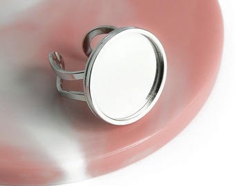Puede incluir: Un anillo de plata con un bisel redondo de vidrio transparente. El anillo está sobre un fondo rosa y blanco.