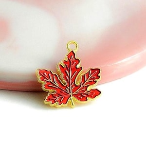 Può includere: Un ciondolo a forma di foglia d'acero rossa con bordi dorati. Il ciondolo è posto su uno sfondo rosa e bianco. La foglia presenta venature dettagliate e un anello per il fissaggio.