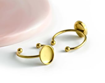 May include: Two gold-coloured adjustable rings with a round bezel setting. The rings have a simple, elegant design with a curved band and small ball accents. A pink, marble-patterned surface is in the background.