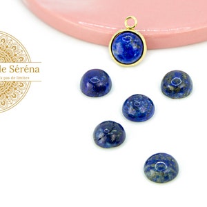 Peut inclure: Six cabochons ronds en lapis-lazuli bleu avec des accents dorés. Les cabochons sont disposés en demi-cercle sur une surface blanche. Un cabochon est attaché à un pendentif en or.