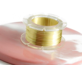 Pode incluir: Um carretel de fio dourado está sobre uma base de resina rosa e branca marmorizada. O fio está enrolado em torno de um carretel de plástico transparente. A base é redonda e tem um acabamento liso e brilhante. Fundo branco.