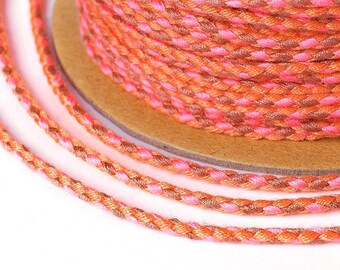May include: A close-up shot of a spool of braided cord in shades of orange, pink, and brown. The cord is tightly wound on a cardboard spool, with several strands extending outwards. The braided texture is clearly visible, showcasing the colour variations.