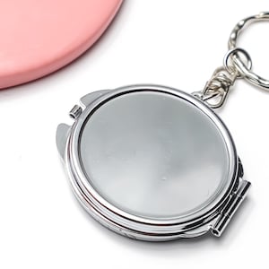 Pode incluir: Um espelho de bolso redondo em metal prateado com porta-chaves. O objeto apresenta uma superfície refletora polida na parte frontal e está preso a um anel metálico circular por um pequeno elo de corrente, servindo como um acessório portátil.