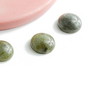 Może przedstawiać: Trzy zielone i szare kaboszony z polerowanym wykończeniem ułożone są na białej powierzchni. Zakrzywiona, różowa ceramiczna miska znajduje się w lewym górnym rogu. Kamienie mają gładki, zaokrąglony kształt.