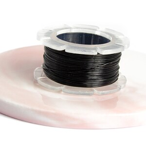 May include: A spool of black wire for jewelry making, on a pink and white marble surface. The spool is clear plastic with a black center.