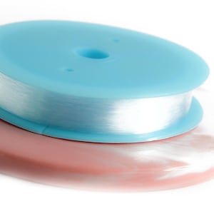 Peut inclure: Une bobine de fil transparent est posée sur un disque en plastique bleu clair, lui-même posé sur un disque plus grand, ovale, brun rosé. Le fil est bien enroulé, et les disques sont lisses et propres.
