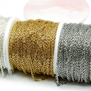 Könnte beinhalten: Drei Spulen mit Ketten in Silber, Gold und Gunmetal-Grau. Die Ketten bestehen aus kleinen, miteinander verbundenen Gliedern und sind um weiße Spulen gewickelt. Der Hintergrund ist weiß mit einem verschwommenen rosa Objekt.