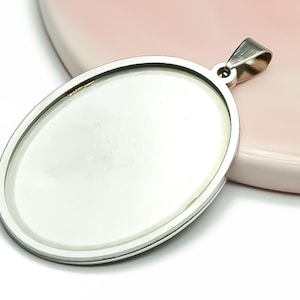 Op de afbeelding: Een zilverkleurige ovale hanger met een lus voor een ketting. De hanger heeft een glad, reflecterend oppervlak en een gepolijste metalen rand. Het ligt tegen een witte en roze achtergrond.
