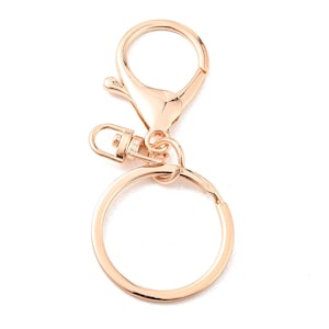 Peut inclure: Un porte-clés de couleur or rose avec un mousqueton, un petit fermoir pivotant rectangulaire et un porte-clés circulaire. Le matériel est en métal et est conçu pour attacher des clés ou d'autres petits objets.
