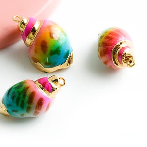 Könnte beinhalten: Drei bunte Muschel-Anhänger mit goldenen Akzenten. Die Muscheln sind in Rosa-, Blau-, Grün- und Gelbtönen bemalt. Die Anhänger sind perfekt, um Schmuck oder Bastelarbeiten eine Prise Verspieltheit zu verleihen.