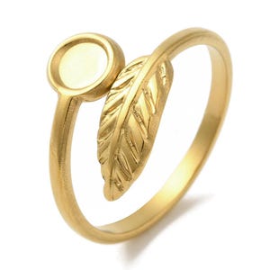 Può includere: Un anello color oro con un design a forma di piuma e un'incastonatura circolare. L'anello presenta una piuma dettagliata che si avvolge attorno al dito, con una piccola incastonatura rotonda sul lato. L'anello è realizzato in metallo lucido color oro.