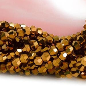 May include: Close-up of a strand of faceted, golden-brown beads. The beads have a metallic sheen and are tightly strung together, creating a textured surface. The background is a soft pink.