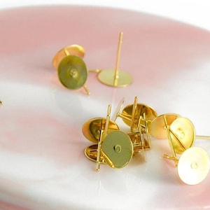 Può includere: Una collezione di perni per orecchini color oro con basi piatte e rotonde e sottili perni. Gli orecchini sono sparsi su una superficie marmorizzata rosa e bianca, creando un'estetica delicata ed elegante.