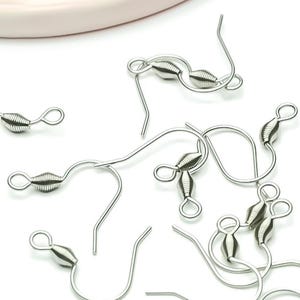 Può includere: Una collezione di ganci per orecchini color argento, sparsi su una superficie bianca. Ogni gancio presenta un design a filo curvo con una perla decorativa e un piccolo anello per attaccare gli orecchini. I ganci sono in metallo lucido.