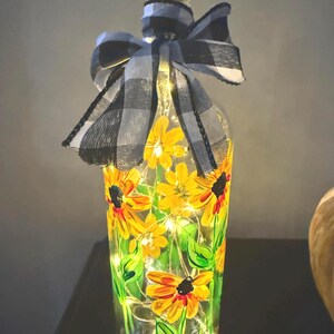 May include: A decorative glass bottle with a cork stopper, hand-painted with yellow sunflowers and green leaves. The bottle is adorned with a black and white checkered bow and illuminated by fairy lights, creating a warm, inviting glow.