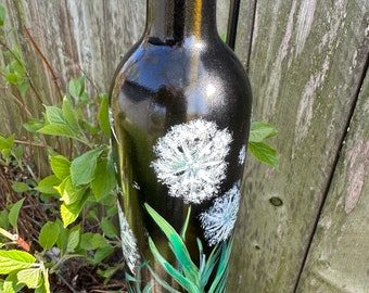 May include: A black glass bottle with a painted design of white dandelions and green leaves. The bottle is hanging from a metal hook.