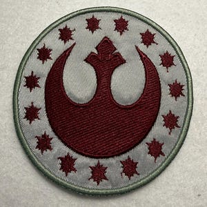 May include: Circular patch featuring a burgundy emblem within a silver field, surrounded by burgundy starbursts and a green border. The emblem is a stylized crescent and crown design. The patch is likely for apparel or accessories.