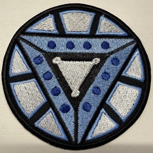 May include: A circular embroidered patch featuring a geometric design with a central white triangle surrounded by light blue and dark blue segments. The pattern includes small dark blue circular accents and is outlined in black thread throughout.