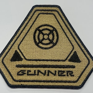 May include: A tan and black embroidered patch with a stylized symbol and the word "GUNNER" in black.